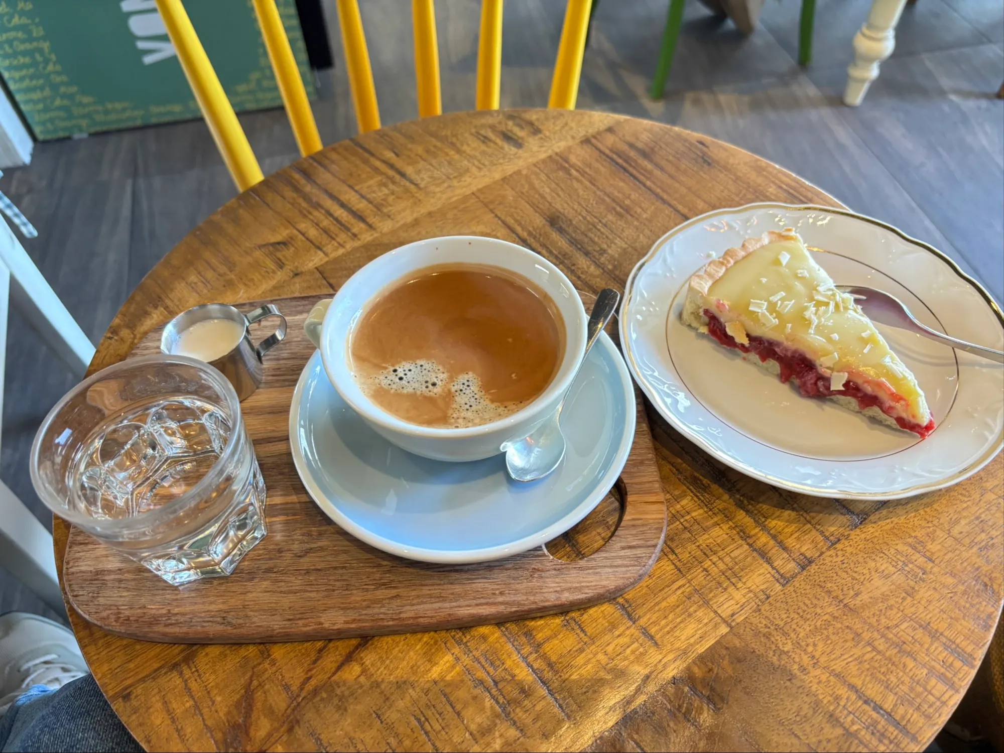 Kaffee in hellblauer Tasse und ein Stück Kirschkuchen auf einem runden Holztisch, dahinter gelbe Stühle