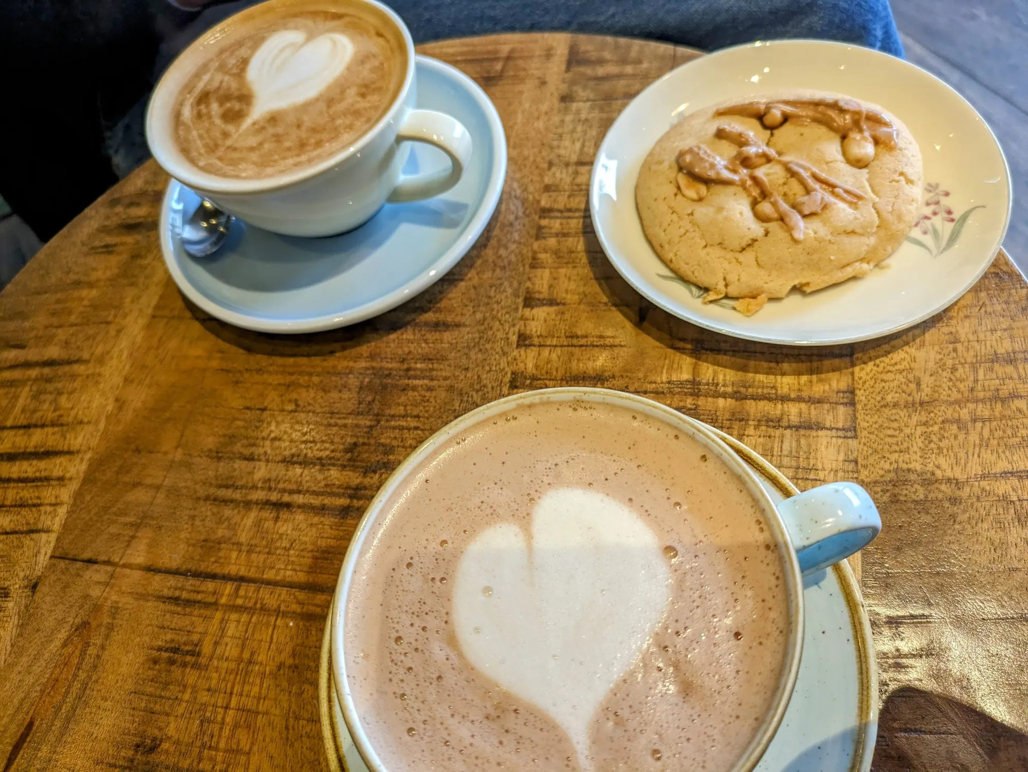 Zwei Milchkaffees mit Herz-Latte-Art in hellblauen Tassen und ein Cookie auf einem runden Holztisch